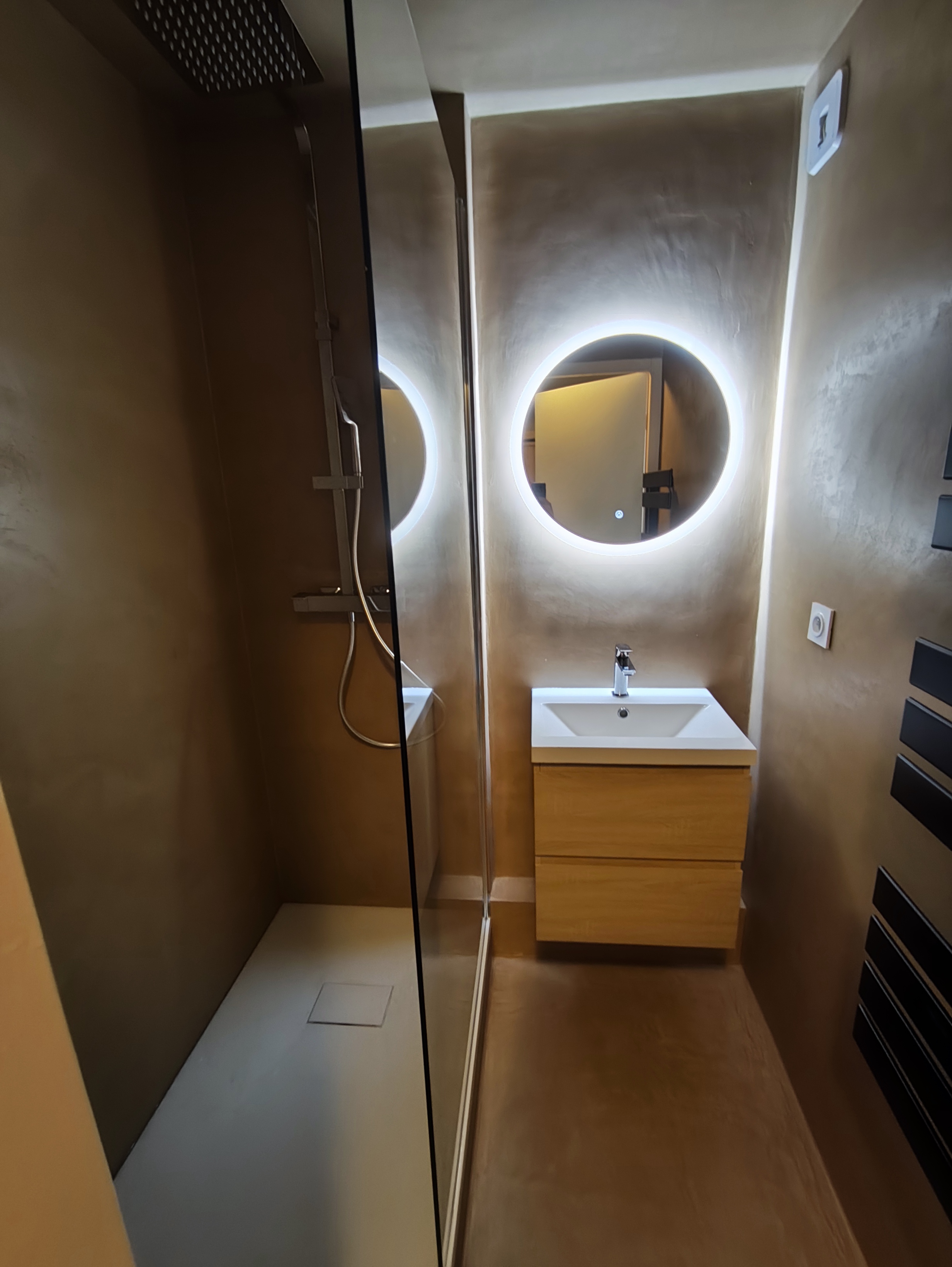 douche vitrée, meuble bois et miroir rond lumineux.